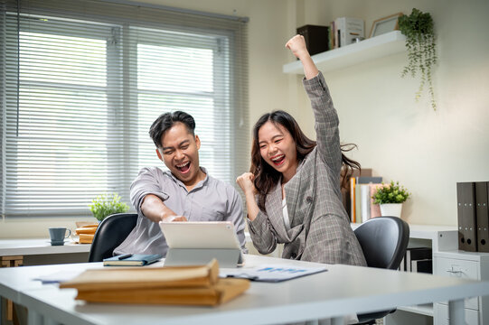 Happy asian businessman and woman coworker raising hand while looking at tablet on table in office. - Powered by Adobe