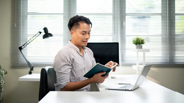Asian businessman or office worker holding and reading a notebook aside laptop sits at working table