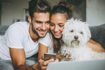 Happy young couple and fluffy white dog smiling while using a smartphone and laptop on a sofa at home, enjoying digital content, cozy domestic lifestyle photography.