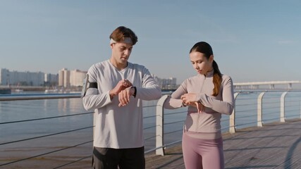 Push in of young male and female athletes in activewear standing on wooden deck by riverside, checking time on their smartwatches, looking at one another and starting morning run - Powered by Adobe
