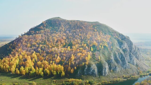 An aerial shot captures the majestic Shihan Juraktau mountain in the South Ural region of Russia. The footage showcases the stunning autumn landscape, with a vibrant forest of yellow and orange trees 