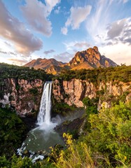 Majestic waterfall cascading down rocky cliffs, surrounded by lush greenery and mountains under a vibrant sunset sky