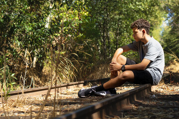 Young Boy Sits Thoughtfully on Abandoned Railway Tracks Surrounded by Overgrown Grass and Trees on a Sunny Day