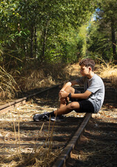 Boy Sitting on Train Tracks in a Forest During Daytime, Lost in Thought Among Tall Grass and Trees