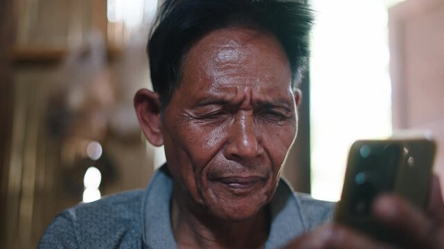 Close-up face and hands of Asian senior man focused on smartphone use inside a traditional lahu house in asia
