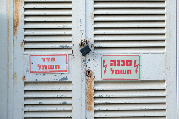 Close-up view of a metal utility door with electrical safety warning signs, featuring a lock and rust marks. Signs indicate high voltage and potential danger in Hebrew