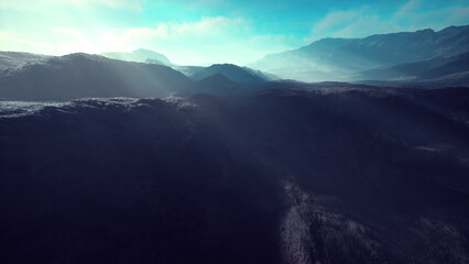 A stunning view of rugged mountains stretching across the horizon, illuminated by soft rays of light breaking through the morning mist. The sky above is a bright blue, creating a serene atmosphere.