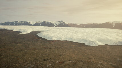 A massive glacier extends over rugged land, creating a striking contrast. In the background, snow capped mountains bask in the warm hues of the setting sun, showcasing natures beauty.