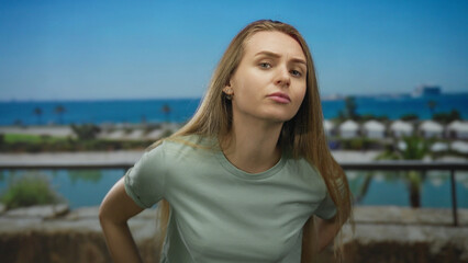 Woman standing outdoors at a port with boats in the background and vibrant blue sea under a clear sky, exuding a thoughtful expression in a casual setting.