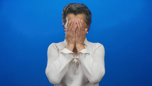 Senior woman with grey hair expresses surprise with wide-eyed hands-on-face gesture against a vibrant blue background in a series of emotional and expressive facial reactions.