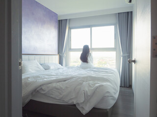 Young woman relaxing in bed with white bedding, smiling in soft morning light by the window