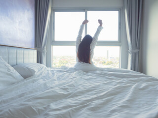 Young woman relaxing in bed with white bedding, smiling in soft morning light by the window