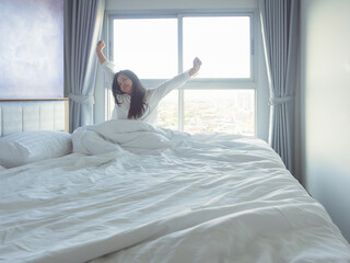 Young woman relaxing in bed with white bedding, smiling in soft morning light by the window