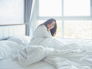Young woman relaxing in bed with white bedding, smiling in soft morning light by the window