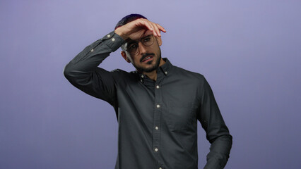 Young hispanic man with glasses posing against a purple background, wearing a dark shirt, looking expressive and thoughtful.