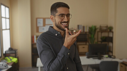 Young man using smartphone in modern office setting, displaying cheerful expression with plants in background indicating a casual professional environment.