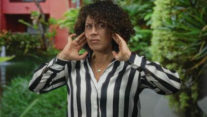 Woman in striped shirt grimaces while covering ears with hands in lush green forest clearing;...