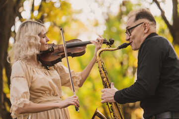 Beautiful blonde violinist and saxophonist perform melodies in autumn park. Fantastic romantic...