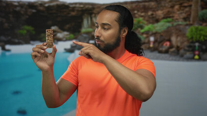 Man holding granola bar points finger to bar at poolside resort hotel deck outdoors while posing;...