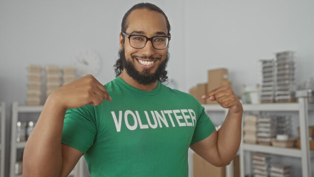 Man points finger to volunteer shirt and smiles with folded arms among packed shelves in a donation center building; community service pride.