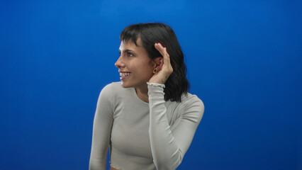 Young hispanic woman cups ear in bright studio with vivid blue wall showing listening gesture;...