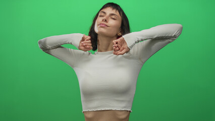 Young hispanic woman yawns with hand covering mouth while stretching arms overhead in green studio wearing crop top; fatigue.