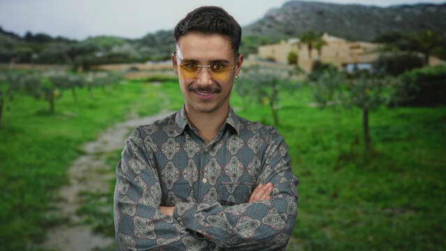 Young hispanic man standing confidently in a lush park landscape, smiling with arms crossed, wearing stylish glasses and a patterned shirt, conveying a relaxed outdoor vibe.