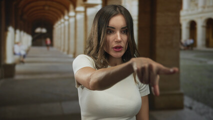 Young hispanic woman points finger to arched corridor building on cobblestone street in old town; curiosity.
