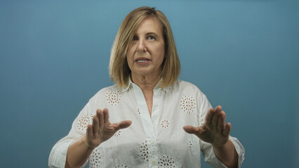 Woman in white blouse holds hands forward with palms facing camera in blue studio; determination.