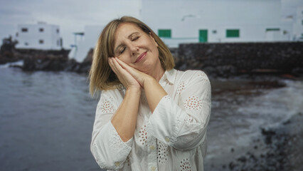 Fototapeta premium Woman places cheek on hands in a sleeping gesture by the seaside with closed eyes and relaxed posture; rest calm serenity relaxation.