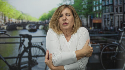 Woman shivering and hugging her arms for warmth on a canal street with blurred bicycles behind her; cold discomfort chill unease.