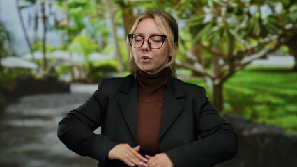 Woman meditating outdoors in a park setting amidst lush greenery, wearing glasses and a dark jacket, exuding calm and focus during a quiet moment of serenity.