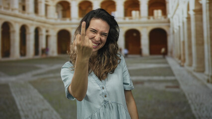 Woman showing middle finger in a sunlit building courtyard with arched corridors and worn stone...