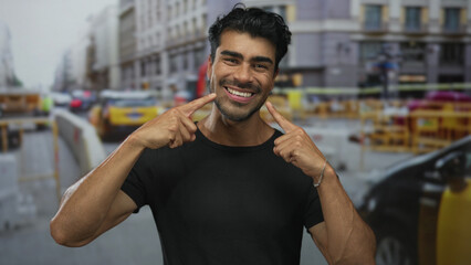Young hispanic man pointing finger to cheeks and smiling broadly among yellow taxis on a busy city street; happiness.