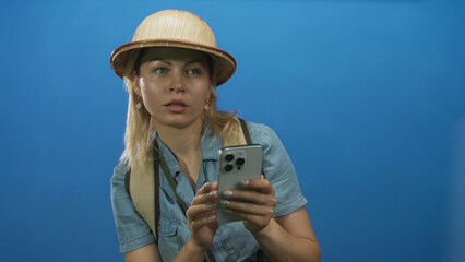 Young blonde tourist woman wearing safari hat and backpack taps smartphone with fingers in blue...