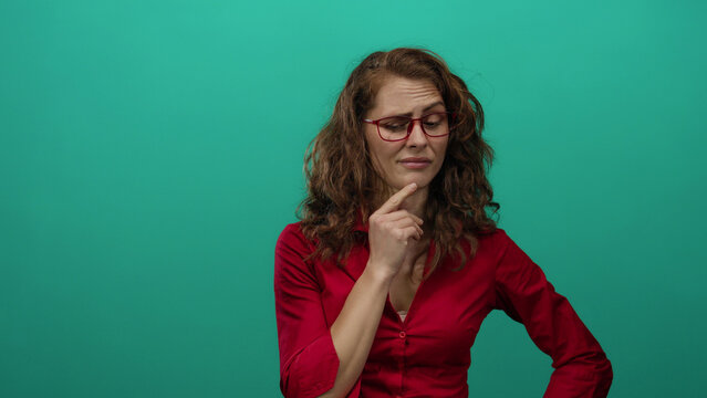 Woman pondering with hand on chin wearing glasses and red shirt against green wall contemplating decision.