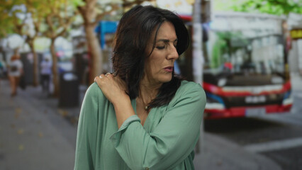 Woman holding shoulder in a pain gesture on a busy urban street beside a red bus and scattered leaves in daylight; pain.