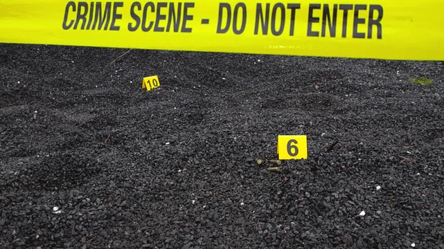 Yellow police line or crime scene forensic investigation tape in front of a dark gravel outdoors area where Crime scene markers are next to evidence like high caliber bullet casing and ammunition.