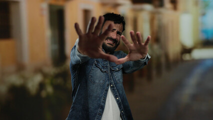 Hispanic man with beard stands outdoors on street appearing scared with hands stretched forward in...