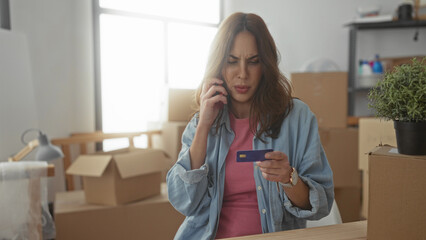 Woman talking on phone while holding credit card in new home with moving boxes around suggests a busy hispanic female managing finances indoors.