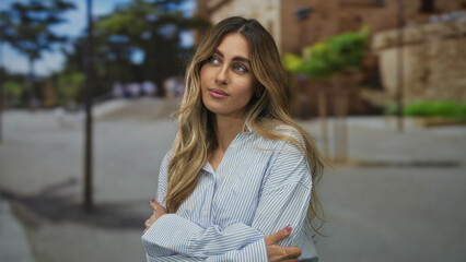 Young blonde woman quietly crosses arms in plaza by stone wall; rejection, doubt, skepticism,...