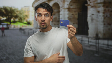 Young hispanic man in white shirt holding credit card near roman coliseum outdoors, looking...
