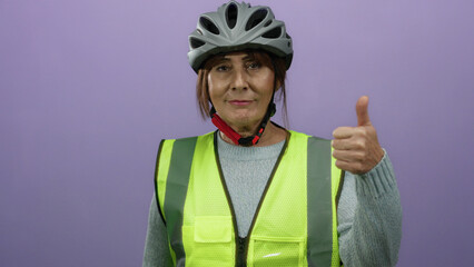 Senior hispanic woman wearing safety vest and helmet stands against a purple wall, giving both thumbs up and down, suggesting mixed feelings or contrasting opinions.