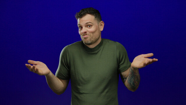 Young man with tattooed arm in green shirt shrugging and expressing confusion against blue background