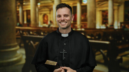 Priest smiling confidently indoors in a church setting, wearing black robe and cross necklace while...