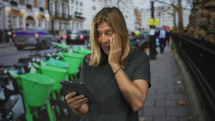 Blond man touches cheek reading tablet on busy urban street amid green parked scooters and cars;...
