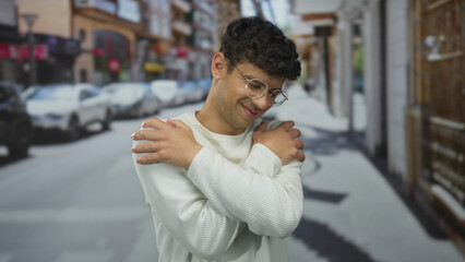 Young hispanic man wearing glasses hugs his shoulders on a street with blurred cars in view;...