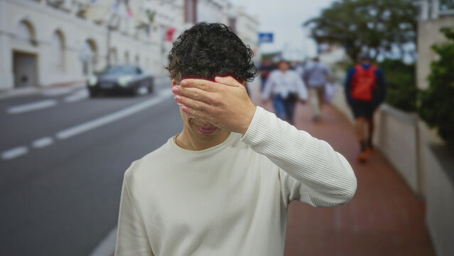 Man covers eyes with hand while standing among blurred pedestrians on busy city street; embarrassment.
