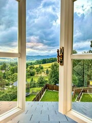 An open window overlooking a serene landscape, green fields and blue skies visible through the frame, inviting fresh air and nature indoors © Diliara