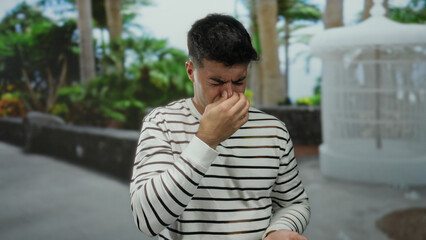 Young man outdoors in a city setting with a pained expression pinching his nose surrounded by lush...